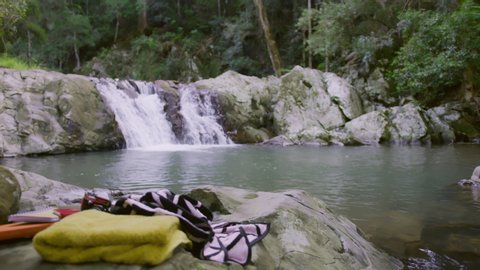 Two beautiful young women undress to bikinis as they are about to swim in a clear pool in a stream with rocky banks with a waterfall in the background and a with towels and a camera in the foreground,
