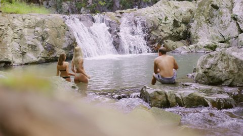 Two beautiful young women swim in a clear pool in a stream with rocky banks with a waterfall in the background as their friend watches, in an Australian forest. Wide shot, in 4K on a RED camera.