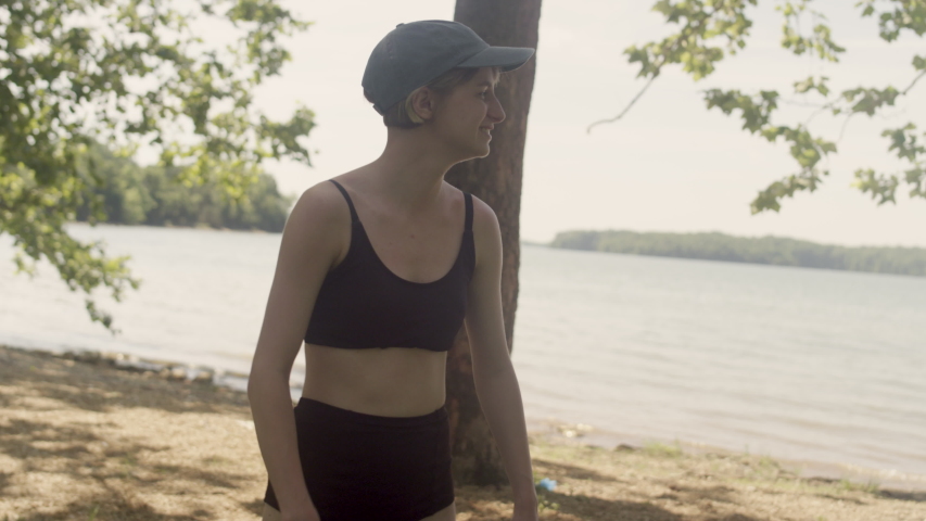 A young woman in a bathing suit and hat points and laughs next to a lake