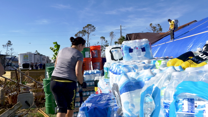 Mexico Beach , Florida / United States - 10 17 2018: A woman hurricane relief volunteer stacks bottled water while a man fixes a roof covered in blue plastic tarp in the background after Hurricane Mic