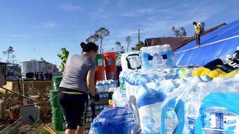 Mexico Beach , Florida / United States - 10 17 2018: A woman hurricane relief volunteer stacks bottled water while a man fixes a roof covered in blue plastic tarp in the background after Hurricane Mic