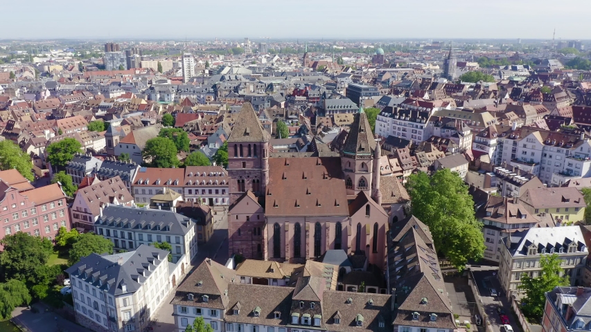 Strasbourg, France. The Protestant Church of Saint-Thomas rises prominently at the center of the frame, its twin towers and broad red-tiled roof dominating the historic cityscape. Built as a five-naved Gothic hall church and completed in 1521, it stands as the only hall church of its kind in the Alsace region, earning it the nickname "Protestant Cathedral." The camera circles the landmark in a smooth aerial orbit, revealing the church