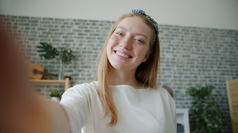 POV portrait of happy girl taking selfie holding camera at home posing touching hair and face. Modern technology, lifestyle and joyful people concept.