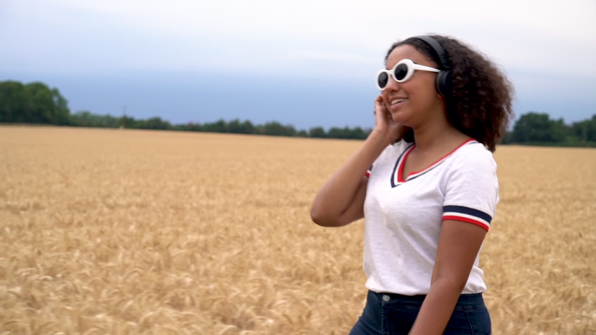 Slow motion tracking video biracial African American girl teenager young woman walking in field, wearing a white t-shirt and sunglasses listening to music on cell phone and wireless headphones