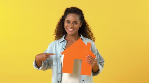 Young African American woman looking at and pointing to house cutout model while acting surprised against yellow background