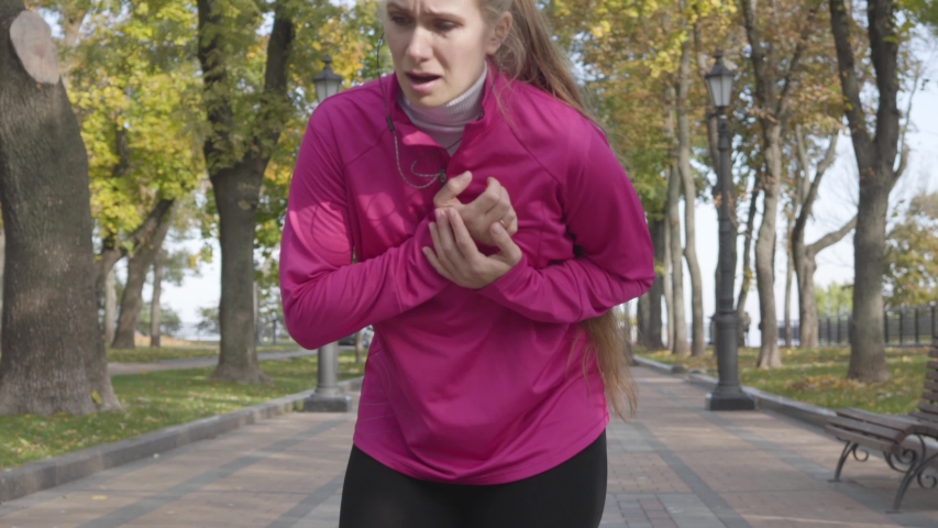 Close-up of an attractive Caucasian woman in pink sportswear having chest pain. Female runner feeling bad during the morning training in the autumn park. Sports and healthcare concept.
