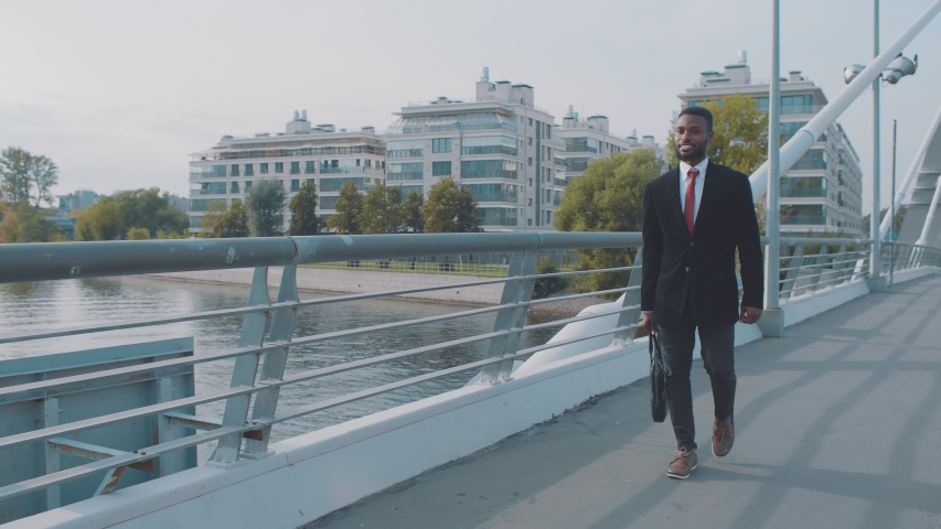 Confident young african american smiling businessman with briefcase walking on modern pedestrian bridge in rich disctrict of city outdoors. Happy bearded black man on background of modern buildings