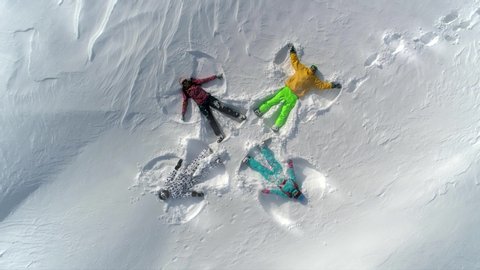 AERIAL TOP UP: a group of young people in bright ski suits depict snow angels lying on the fresh snow in the form of a cross. four winter angels happy and energetic with splashes of snow. SLOW MOTION