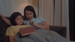 Young Asian family and daughter happy using tablet at home. Korean mother relax with little girl watching movie lying on sofa before going to sleep in living room at house in night.