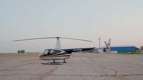 Drone flying left to right. Aerial view of a helicopter standing on air base at sunset time.The main rotor blades are rotating slowly 