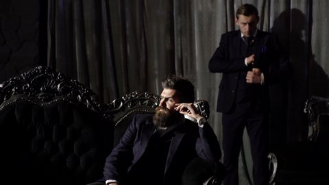 Bearded man with serious face and his security guard in classic interior. Millionaire in elegant suit sits on luxurious sofa. Brutal man with his assistant. Affluent, security, secret service concept