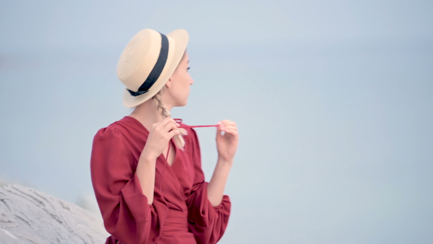 Portrait attractive caucasian young woman in a red dress and a straw hat with pigtails turns around sitting on the seashore smiling looking at the camera against the sea in defocus