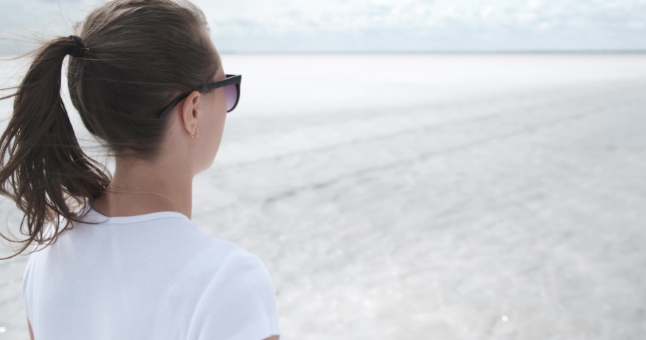 Girl in slow motion walking along beach, clear water waves, girl smiles, looks around, great summer weather, distant resort, salt lakes, medical tourism, the opportunity to improve health, salt sea