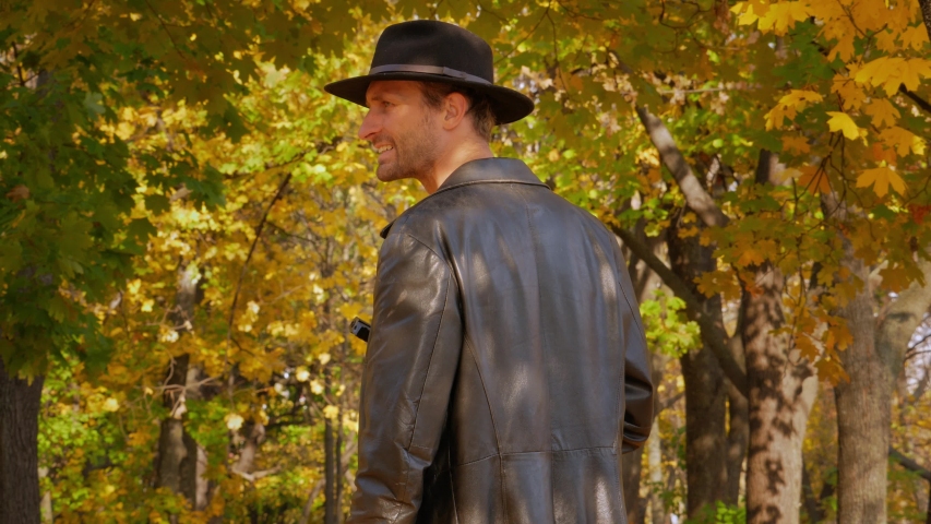 Dangerous man in a black cloak and hat stands with a gun in the autumn forest and looks around