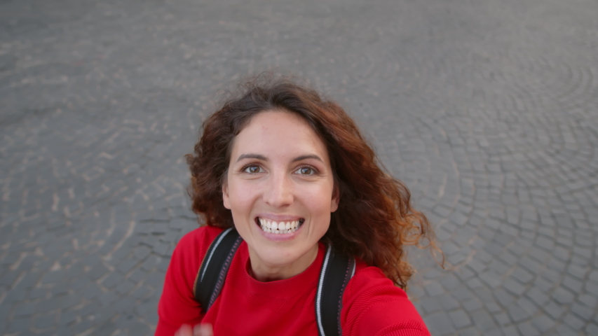 Portrait shot of happy young woman with curly hair laughing and filming herself outdoors. She is waving and gesturing