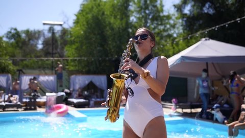 Attractive saxophonist lady is playing on saxophone near pool at beach club. Pretty sax girl musician in hot white bikini dances and plays on weekend party onhot summer day.
