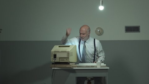Angry mature businessman sitting at desk and hitting his outdated old computer