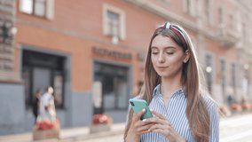 Pretty Young Girl with Brown Hair and Stylish Headband Wearing Striped Shirt Having a Video Call on her Smartphone Smiling and Waving Hand Walking at City Street