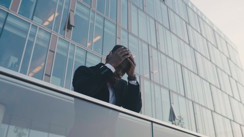 Portrait of upset desperate man with headache against office center as background. Black guy in formal suit getting bad news, touching head with hands feeling frustrated