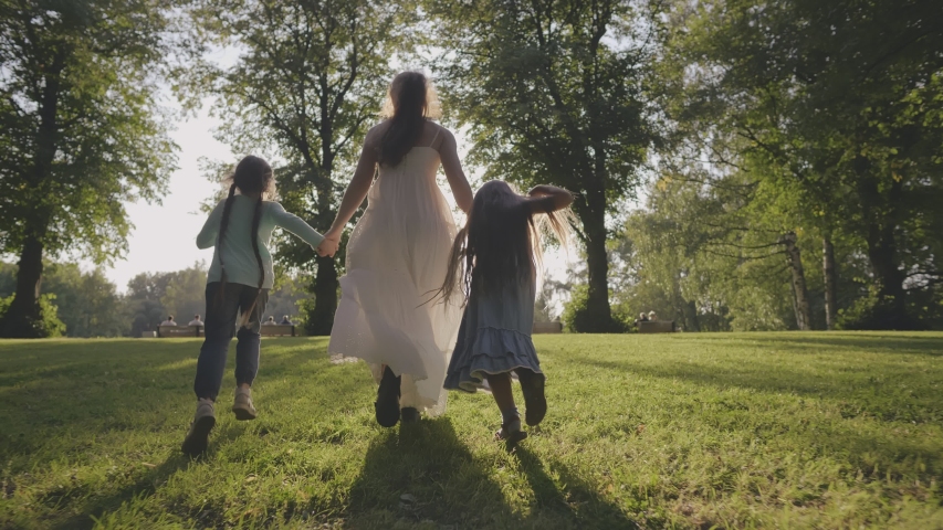 Young mother with siblings daughters together running towards sun having fun in city park garden. Family run on green grass at background of trees during summer sunset. Motherhood childhood concept | Shutterstock HD Video #1039232024