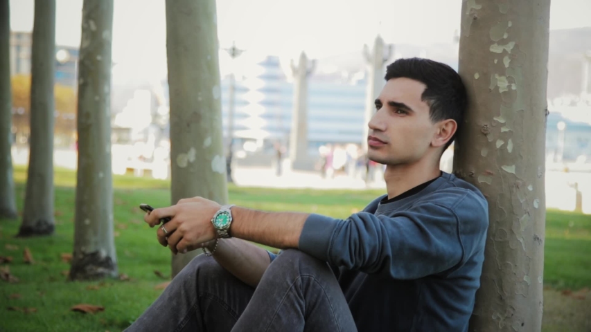 Portrait of young man sitting near tree and thinking in warm autimn day, In slow motion