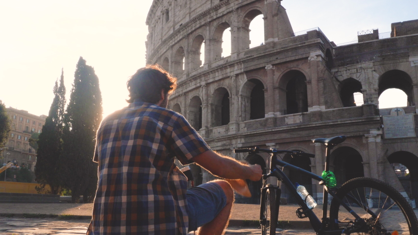 Happy young man tourist wearing shirt and hat with bike sitting and taking pictures with vintage camera at colosseum in Rome, Italy at sunrise.