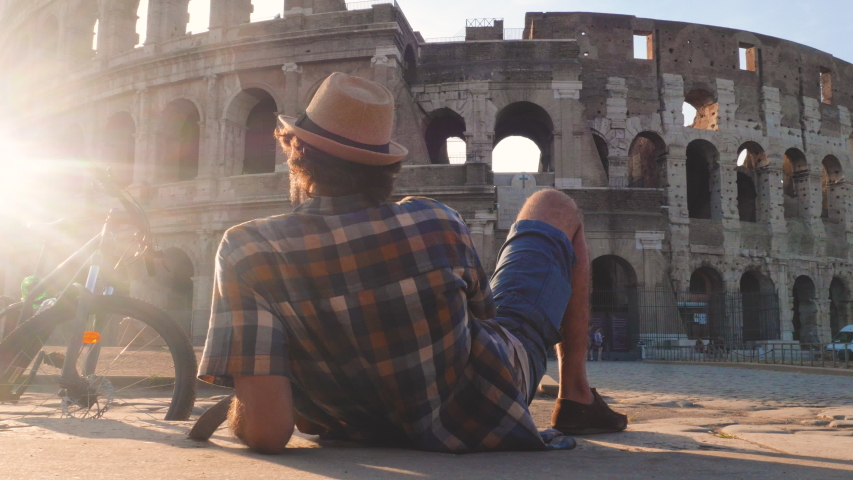 Happy young man tourist wearing shirt and hat with bike sitting and taking selfies with vintage camera at colosseum in Rome, Italy at sunrise.
