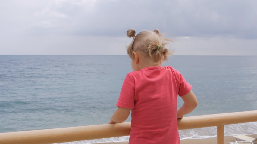 A little girl stands on the waterfront overlooking the sea, leaning on the fence.