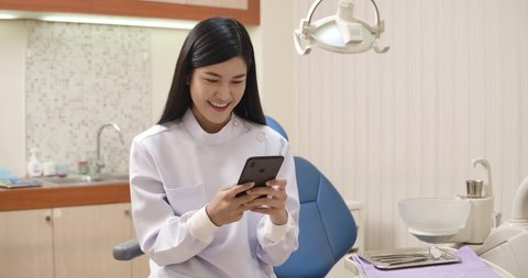 Close-up portrait of young Asian woman dentist using mobile phone on a chair in a dental clinic while waiting for patient appointment, young woman smiling while browsing internet.