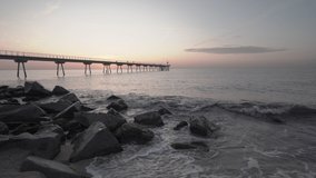 4K clip of an early morning sunrise behind a huge concrete bridge in the mediterranean sea with rocks and crashing waves in the foreground and people walking and running at the beach