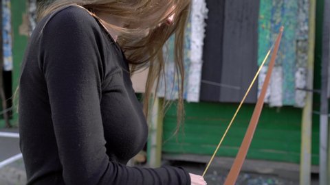 A girl shoots from a bow at targets. Archery sports, outdoor training. Wooden bow and arrow, accuracy, hitting the target. Evening light. Female focusing to the target. 