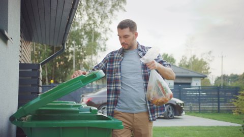 Caucasian Man is Walking Outside His House in Order to Take Out Two Plastic Bags of Trash. One Garbage Bag is Sorted into Biological Food Waste, Other is Thrown into Recyclable Bottles Garbage Bin.