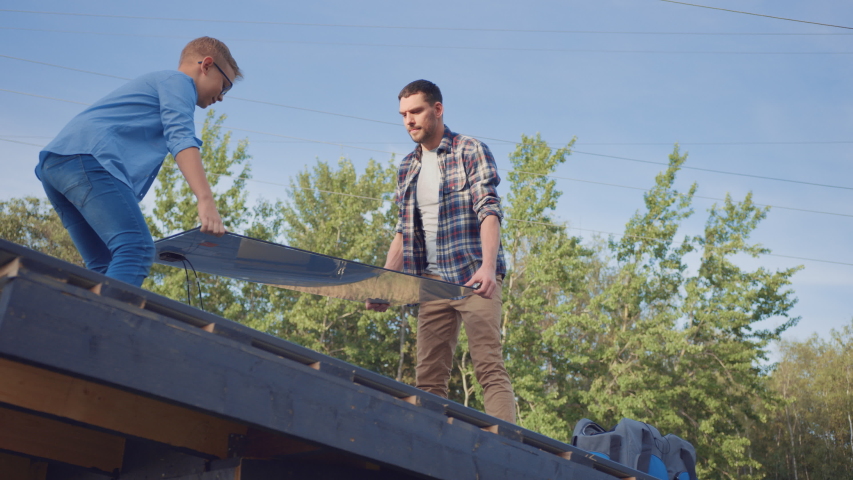 Father and Son Installing Solar Panels to a Metal Basis. They are Holding the Panels on a House Roof on a Sunny Day. Concept of Ecological Renewable Energy at Home and Quality Family Time. Royalty-Free Stock Footage #1039252421