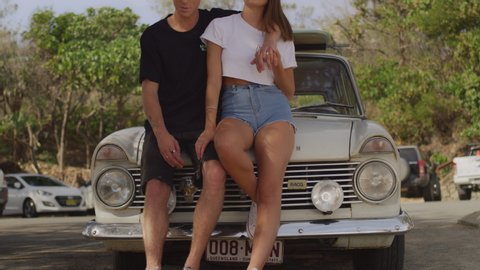 Young couple leaning against classic station wagon with surfboards on beach looking at ocean with surfers in the background.Medium close on 4k RED camera.