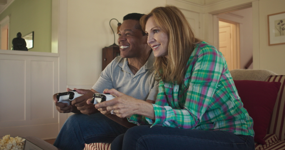Happy older couple playing video games together on living room couch. African American and Caucasian husband and wife using console controllers to play videogame. Slow motion 4k handheld