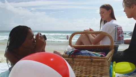 Friends hanging out on a beautiful beach taking photographs and having a picnic, swimmers and surfers in the background. Medium close on 4k RED camera.