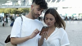 Front view of smiling young couple looking at smartphone. Cheerful people talking and using phone while standing on square. Technology concept