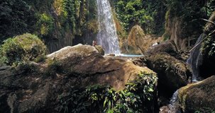 Drone shot revealing stunning waterfall, camera moving forward in slow motion. Aerial view of waterfalls, turquoise colored water and Filipino girl sitting on rock as subject.