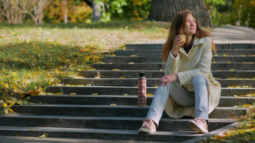 Young woman holding in hands cup of hot tea, sitting on stairs in outdoors autumn park.