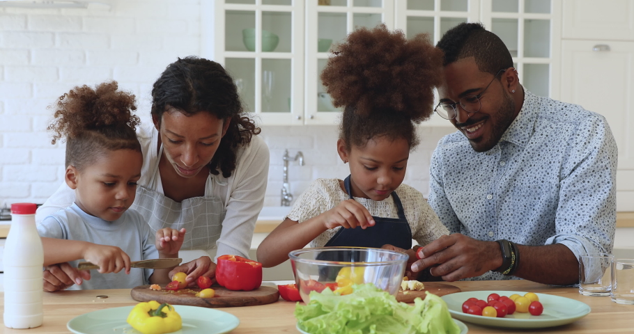 Cute little african american boy and girl help parents cutting fresh vegetables tomatoes pepper for healthy salad, happy mixed race family mom dad and kids girls cooking together in modern kitchen Royalty-Free Stock Footage #1039317596