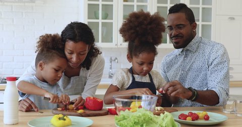 Cute little african american boy and girl help parents cutting fresh vegetables tomatoes pepper for healthy salad, happy mixed race family mom dad and kids girls cooking together in modern kitchen