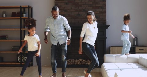 Joyful active african family dancing jumping in modern living room interior, carefree happy mixed race young parents mom dad with cute little kids children boy and girl having fun together at home