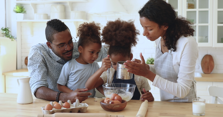 Cute little african american daughter holding whisk helping happy mixed race family preparing dough, smiling parents mom dad and kids children siblings baking cake pastry together in modern kitchen Royalty-Free Stock Footage #1039317614