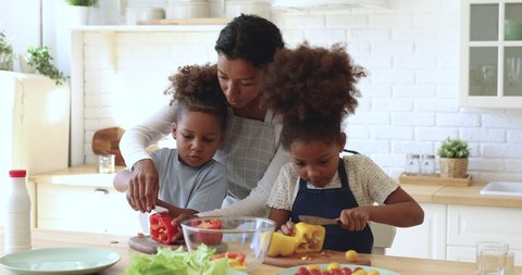 Smiling young multiracial mum teaching cute kids to cook. Cutting fresh organic vegetable salad in a kitchen. Happy family. Little children helping mom prepare healthy food together at home.