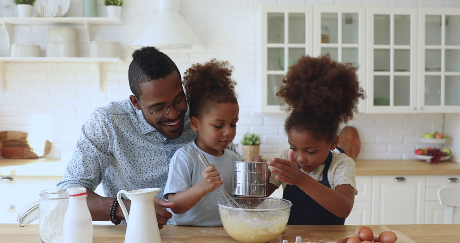 Cute african american family young father dad with mixed race little children kids siblings preparing dough together whisking flour and eggs having fun cooking baking cake cookies in modern kitchen Royalty-Free Stock Footage #1039317629