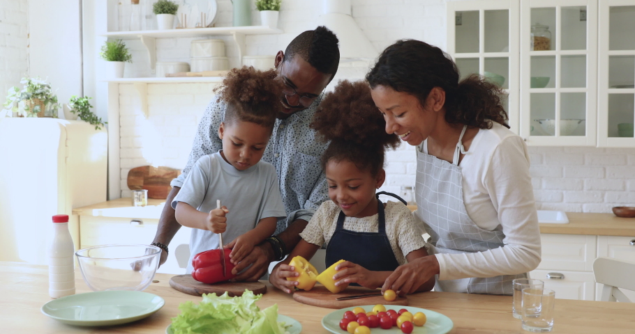 Happy african family mom dad and small cute kids cut fresh vegetable salad cooking together in modern kitchen, mixed race parents teach little children son daughter help prepare healthy food at home Royalty-Free Stock Footage #1039317647