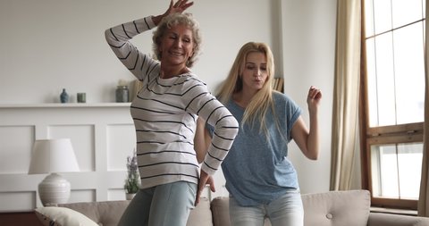 Happy carefree two age generation women family young grown adult daughter and healthy active old senior mature mother listening music dancing together laughing having fun at home in living room