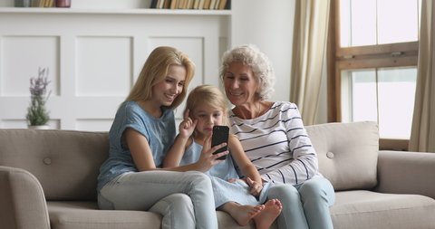 Happy three 3 age multi generation women family old grandmother young adult mother and child girl granddaughter have fun use smart phone funny video face app take selfie together at home sit on sofa
