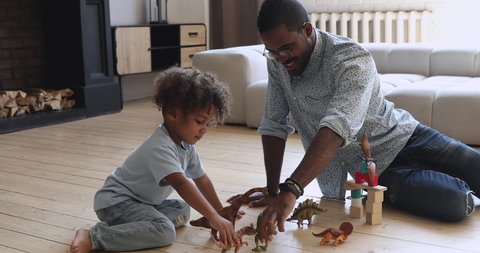 Happy african american family young dad babysitter and cute small kid son pretending dinosaurs battle sit on floor at home, father playing game with little child boy holding toys having fun together