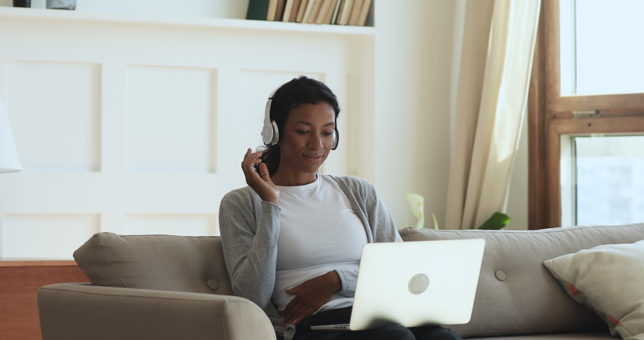 Happy young african woman wear wireless headphones make video call online chat look at laptop screen sit on sofa, smiling girl talk to webcam communicate by internet computer conference app at home Royalty-Free Stock Footage #1039317809